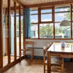 Dining room with wooden table, chairs, and large windows. View of garden and adjacent buildings.