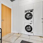 Waschmaschine und Trockner in weißer Einbauküche mit Holztür und schwarzer Garderobe.