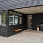 Terrasse mit Grill, Waschmaschine und Holzbänkchen an schwarzem Holzhaus.