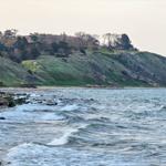 Beach with rocks and waves, behind green hills with trees and a red house.