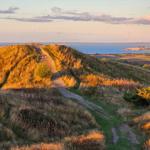 Path leads over grassy hill to coast with view of fields and sea.
