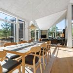 Dining area with wooden table and chairs. Large windows show forest. Living area with sofa and TV.