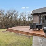 Wooden terrace with seating and grill beside house. Lawn and trees in background.