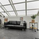 Sofa und Sessel unter Glasdach mit Pflanze und Wandbildern.
