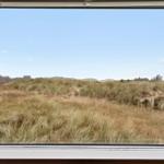 Durch das Fenster blickt man auf eine Graslandschaft mit Dünen und Häusern im Hintergrund.