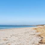 Strand mit Sand, trockener Vegetation und Blick aufs Meer und Ferienhäuser.