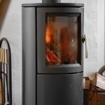 Schwarzer Kaminofen mit brennendem Holz, daneben Holzstapel und Feuerzeugständer.