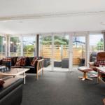 Wohnraum mit Sofa, Sessel und Tischen. Durchgehende Glasfront zum Garten.
