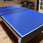 Blue ping pong table with net in a room with wooden floor and sofa.