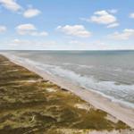 Aerial view of a long sandy beach with dunes and sea under a partly cloudy sky.