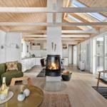 Wohnzimmer mit Kamin, Holzdecke und großem Fenster. Küche im Hintergrund.