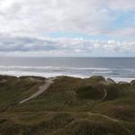 Meer mit Dünen und Wanderpfaden unter bewölktem Himmel.