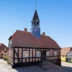 Traditionelles Fachwerkhaus mit Turm und Uhr, umgeben von historischen Gebäuden auf Kopfsteinpflaster.