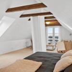 Schlafzimmer mit Dachschrägen, Holzbalken und Fenster. Bett, Sofa und Lampen sichtbar.