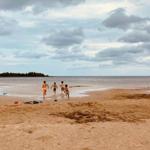 Vier Kinder laufen am Strand entlang, im Hintergrund See und bewölkter Himmel.