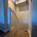 Treppenhaus mit Holzstufen und weißer Brüstung. Deckenleuchte und neutrale Wände.