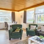 Wohn- und Essbereich mit grünen Sesseln, Sofa und Glas-Tisch. Holzdecke und große Fenster.