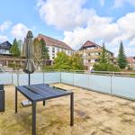 Terrasse mit Tisch, Stuhl und geschlossenem Sonnenschirm. Glasgeländer und Sicht auf Nachbarhäuser.