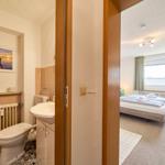 Badezimmer mit Toilette und Waschbecken. Blick in das Schlafzimmer mit Bett und Fenster.