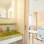 Badezimmer mit grünem Waschbecken und Spiegel. Blick in Schlafzimmer mit Bett und Tisch.