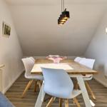 Esszimmer mit Holztisch, weißen Stühlen und schrägen Wänden. Fenster, Pflanze und Deckenleuchte sichtbar.