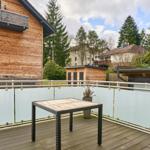 Terrasse mit Tisch, Glasgeländer und Blick auf Holzhaus und Bäume.