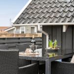 Terrasse mit Tisch und Stühlen, darauf Flasche und Blumen. Hintergrund: Haus mit Dach und Zaun.