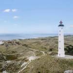 Weißer Leuchtturm auf grünen Dünen mit Blick auf das Meer.