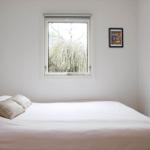 A bed with pillows under a window with a view of trees. A picture hangs on the wall.