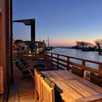 Terrasse mit Tisch und Stühlen an Wasser, Blick auf Hafen und Sonnenuntergang.