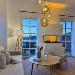 Wohnzimmer mit Sofa, Sessel, Kamin und Holzboden. Fenster und Hängelampe sichtbar.