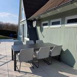 Terrasse mit Tisch und sechs Stühlen vor einem Haus mit grünem Holzverkleidung.