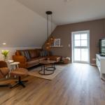 Wohnzimmer mit braunem Sofa, Sessel und Holzboden. Fenster und TV-Schrank sichtbar.