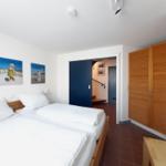 Zimmer mit Doppelbett, Holzschrank und Wandbildern. Tür führt in einen Flur mit Treppe.