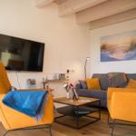 Wohnzimmer mit grauem Sofa, zwei orangefarbenen Sesseln und Fernseher an der Wand.