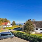 Haus mit Strohdach, Garten und Blick auf Wasser. Parkplatz mit zwei Fahrzeugen.