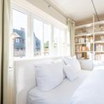Schlafzimmer mit weißem Bett, Bücherregal und Fensterblick auf Dachhäuser.