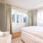 Schlafzimmer mit weißem Bett, Sessel und großen Fenstern.