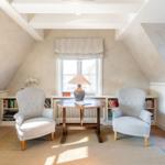 Zwei Sessel und ein Tisch stehen vor einem Fenster in einem Dachzimmer mit Bücherregalen.