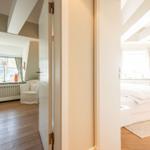 Zimmer mit Bett und Bücherregal. Durchgang zu Wohnbereich mit Fenster und Sessel.