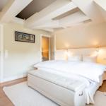 Schlafzimmer mit Doppelbett, Holzboden und weißen Wänden. Deckenbalken und Fenster sichtbar.