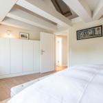 Schlafzimmer mit weißem Bett, Holzboden und Deckenbalken. An der Wand hängt ein Bild.