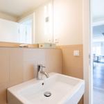 Bad mit Waschbecken, Spiegel und Lichtleisten. Tür führt in Wohnbereich.