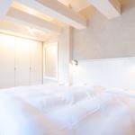 Schlafzimmer mit weißem Bett, Kleiderschrank und Wandlampen unter Holzbalkendecke.