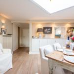 Essbereich mit Holztisch und weißen Stühlen. Hintergrund zeigt Küchenbereich und Wohnbereich.