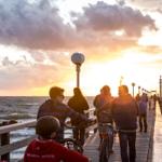 Besucher gehen auf einem Steg entlang, während Sonnenuntergang über Meer sichtbar ist.