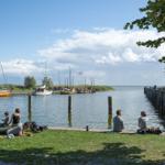 Besucher sitzen am Ufer, Boote liegen im Hafen, Bäume säumen den Weg.