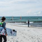 Person mit Windsurfboard am Strand, im Hintergrund Wasser und weitere Surfer.