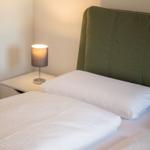 Ein Bett mit weißer Bettwäsche und grünem Kopfteil. Neben dem Bett steht ein Nachttisch mit einer Lampe.