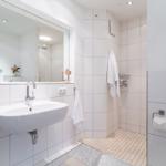 Badezimmer mit Waschbecken, Spiegel und Dusche. Weiße Fliesen und Handtuchhalter.